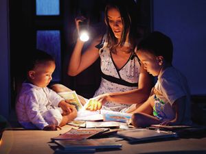 A mother and her two children read storybooks by torchlight on the floor.